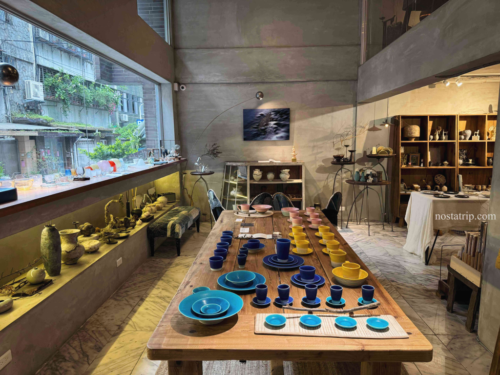 一嶼 Island 室內展陳空間，木桌上整齊擺放藍色、黃色與粉色陶器，窗邊自然採光灑入