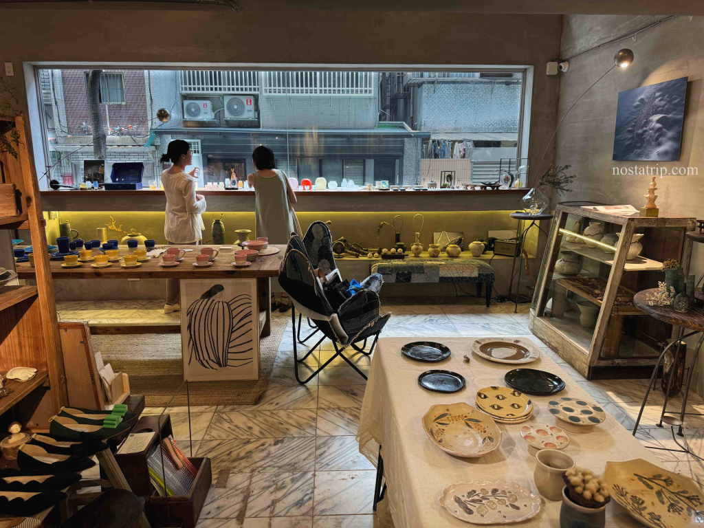 一嶼 Island 店內空間中，兩位女性正於窗邊觀察展示器物，桌面陳列手繪陶盤與彩色陶杯