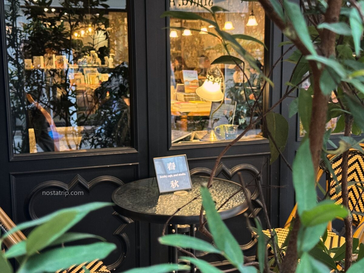 赤峰街春秋書店窗外的圓桌與藤編椅，桌上放著「春秋」立牌，綠葉後方可見窗內暖色檯燈。